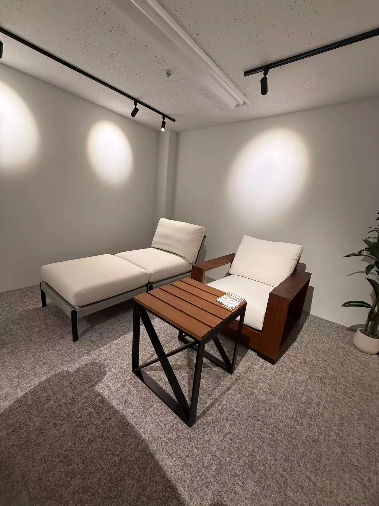A lounge chair with ottoman and a wood-framed armchair, both with white cushions, beside a slatted wooden side table under spotlights